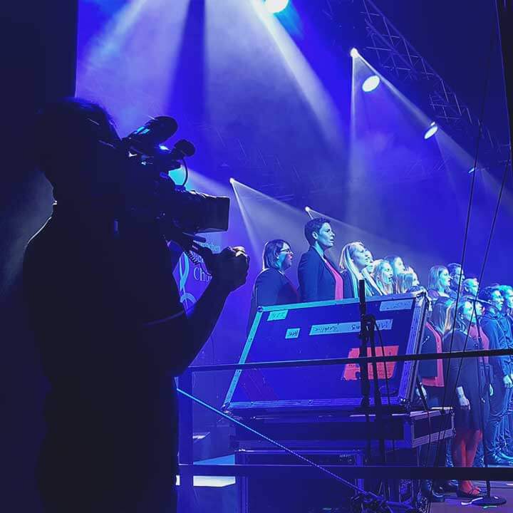 Person filmt mit Kamera einen gemischten Chor auf einer Bühne. Beleuchtung in Blau